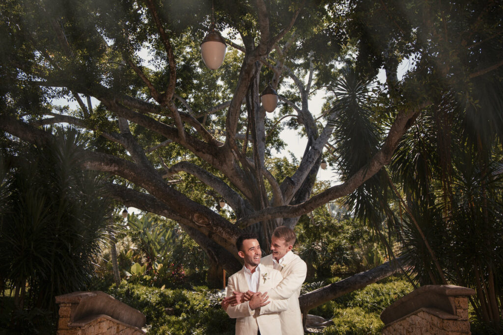 Under the shade of a majestic tree, two grooms in cream suits share a joyful moment, with their hands intertwined, symbolizing their union in a lush garden setting