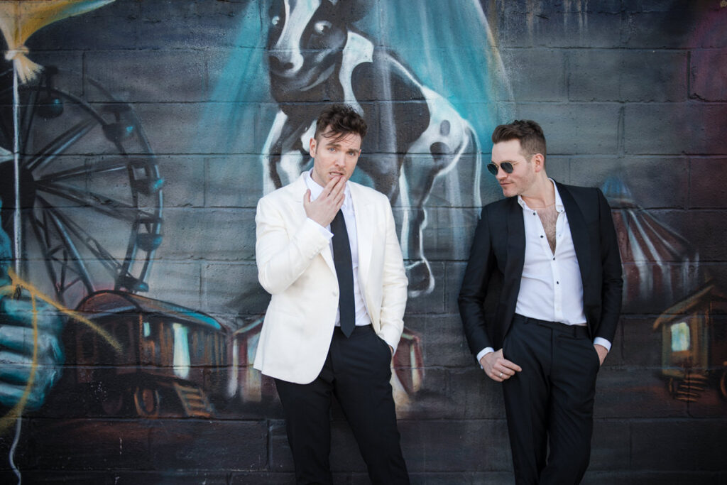 Two grooms in stylish white and black suits pose in front of a vibrant graffiti wall, one with a contemplative expression and the other with a smirk, reflecting a modern and urban wedding theme