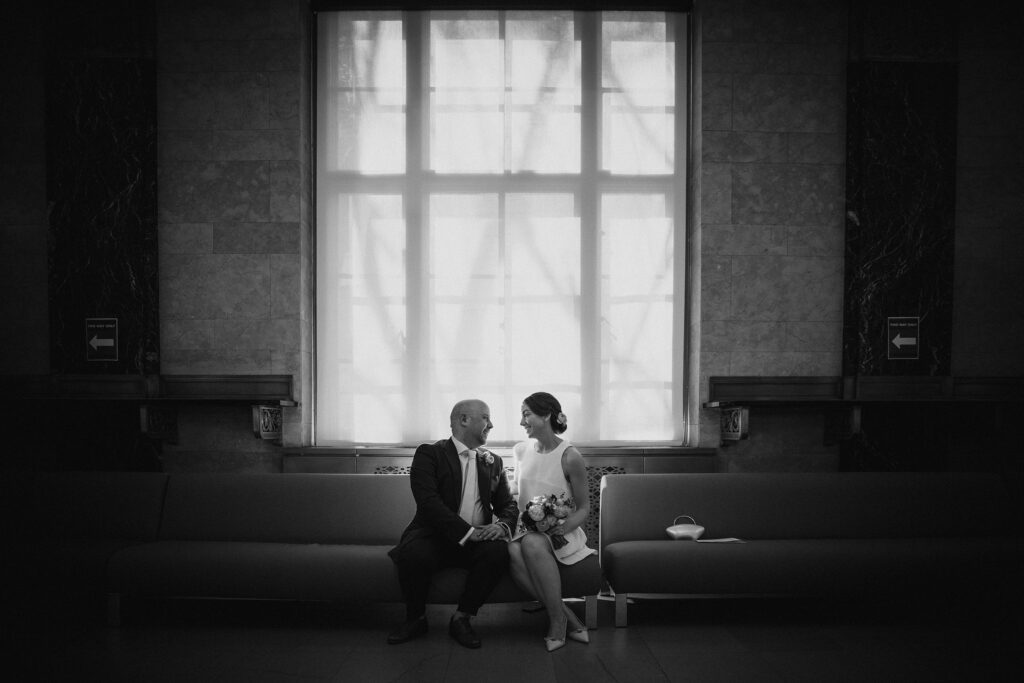 A bride and groom sitting on a couch in front of a window, capturing the essence of their intimate NYC elopement at the marriage bureau.