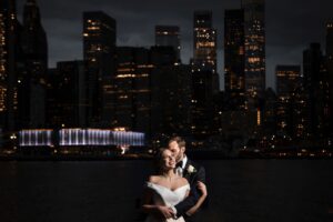 A bride and groom posing in front of a city skyline at night at the River Cafe