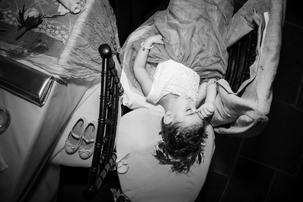 Capturing the power of real moments, a little girl peacefully lays on a chair at a wedding reception without her shoes on.