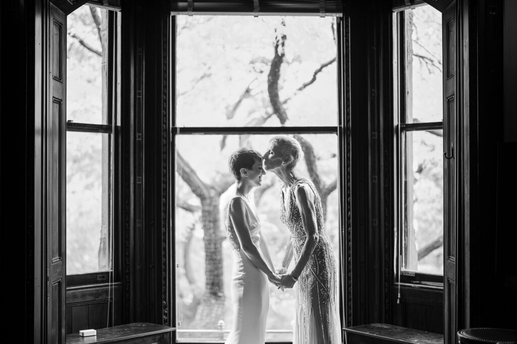 A bride and her mom having a moment while standing in front of a window on a wedding day.