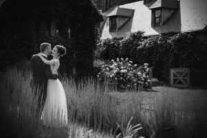 A man and woman sharing a passionate kiss in front of a house during their enchanting Blue Hill at Stone Barn wedding.
