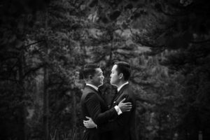 A black and white photo of two men hugging in the woods for their wedding first look.
