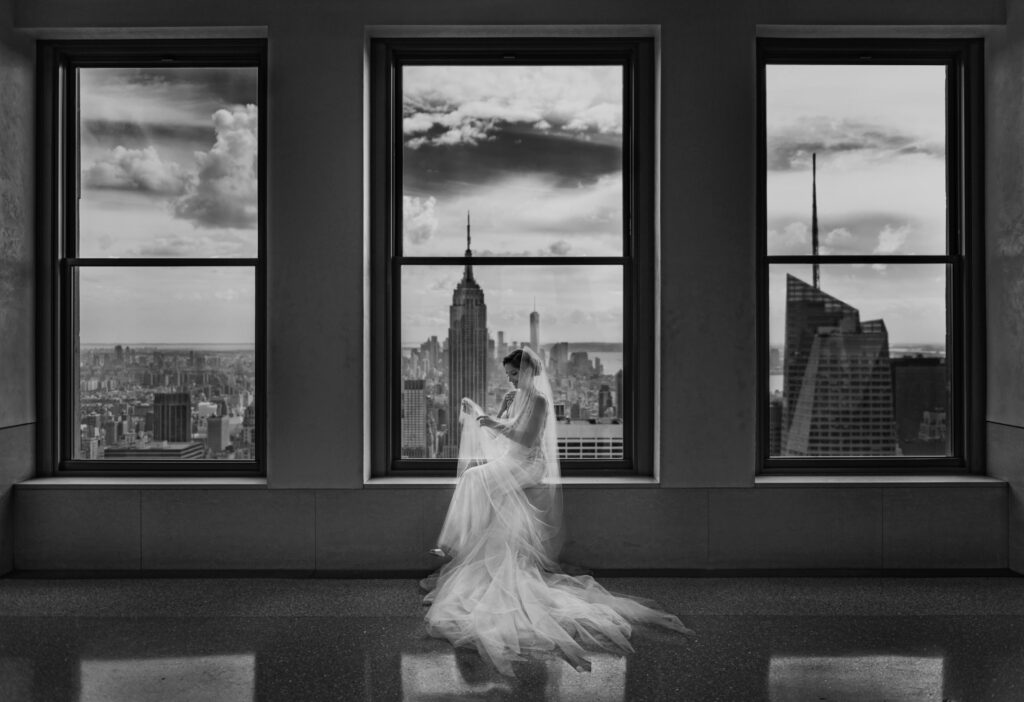 A bride is standing in front of a window overlooking the city during her Top of the Rock wedding.
