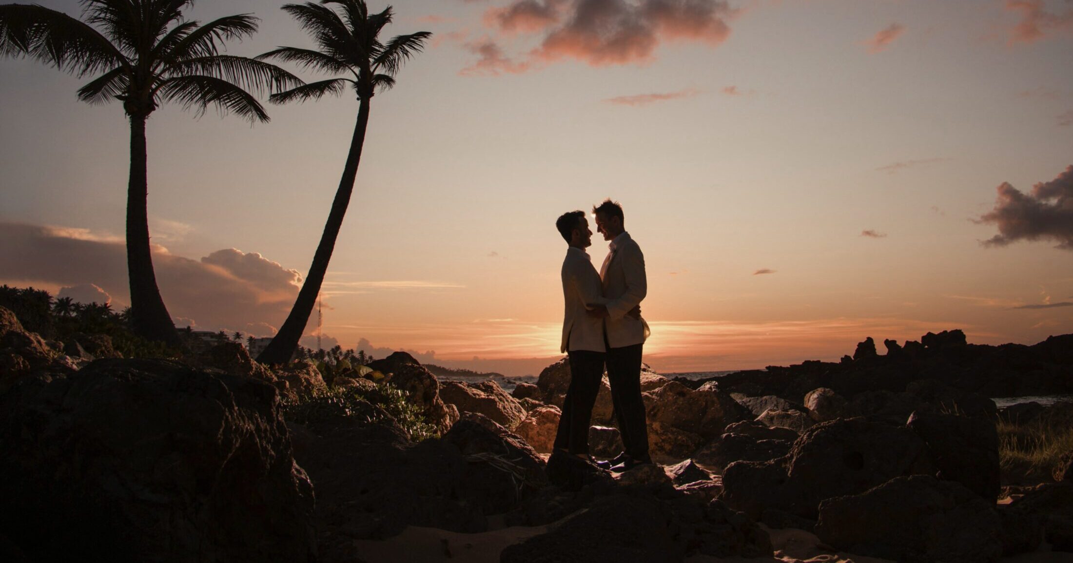 A couple captured during their destination wedding, standing on rocks at sunset with palm trees in the background.