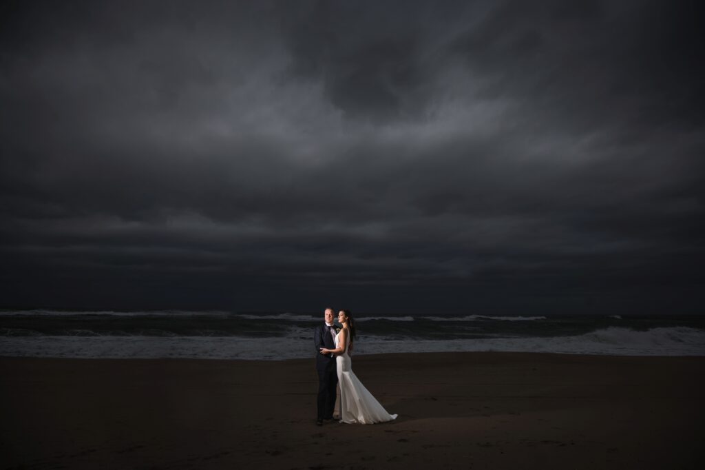A bride and groom standing on the beach at Gurney's Montauk wedding, under a stormy sky.