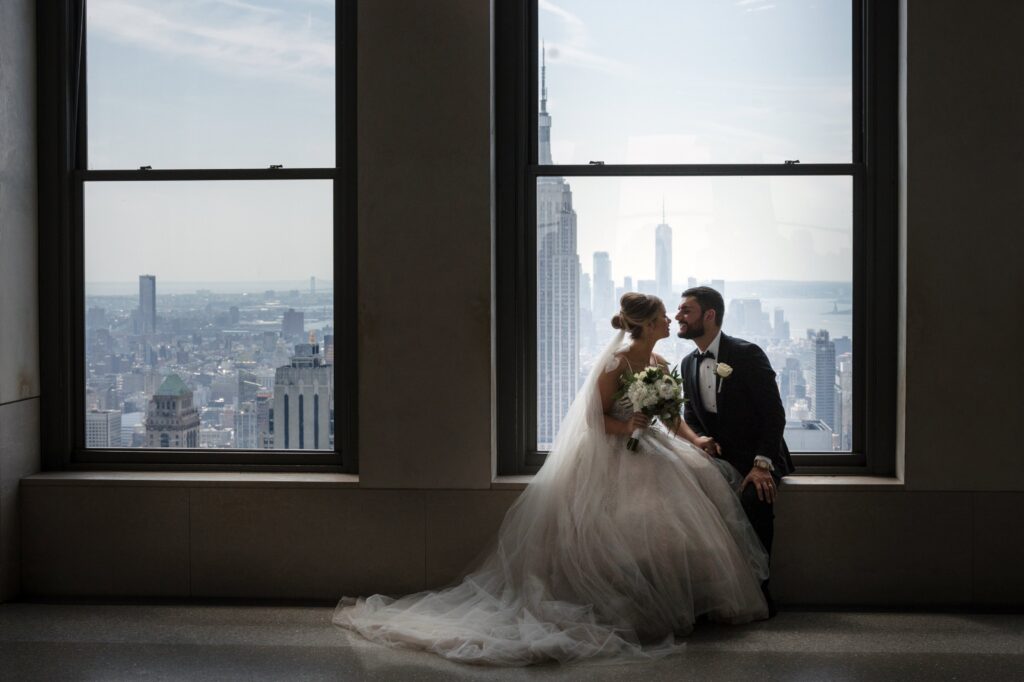 A Top of the Rock wedding couple pose in front of a window overlooking the city.