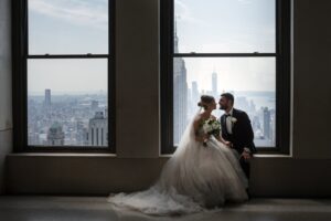 A Top of the Rock wedding couple pose in front of a window overlooking the city.