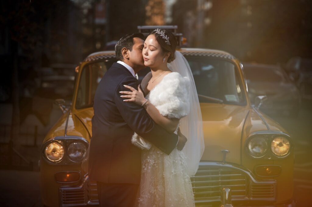A newly married couple share a tender kiss in front of a vintage taxi on their wedding day.