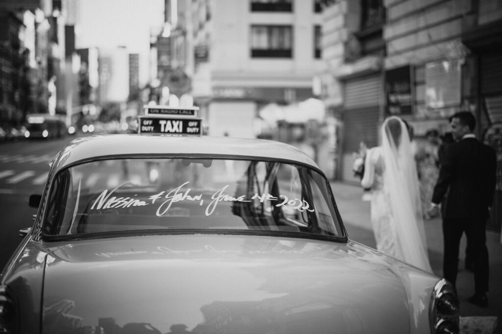 A black and white photo of a bride and groom standing in front of a taxi at their Capitale wedding.