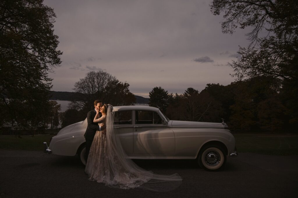 A bride and groom elegantly pose next to a vintage car at dusk during their Lyndhurst Mansion wedding.