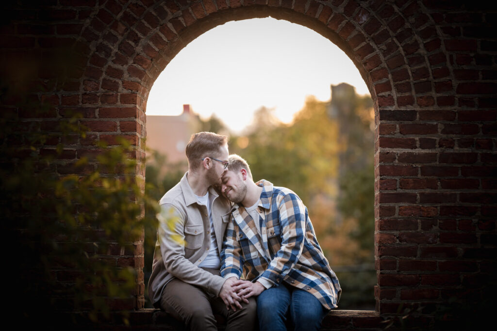 A joyful couple seated in a rustic brick archway, dressed in casual engagement outfits with plaid and neutral colors