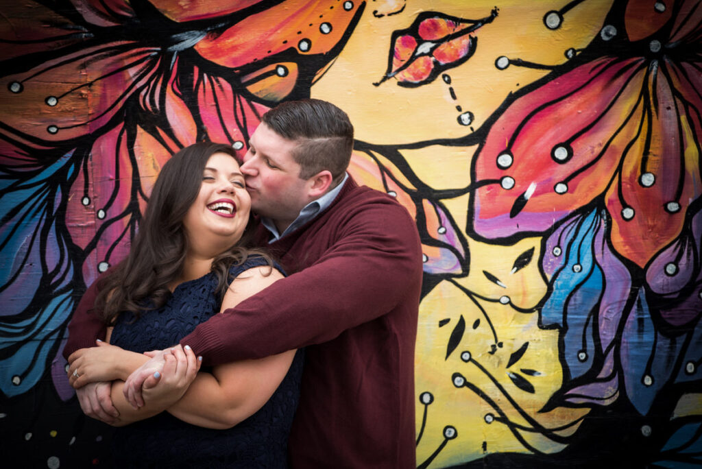 A couple laughing and embracing in front of a colorful mural, showcasing casual engagement outfit ideas with coordinated dark blue tones