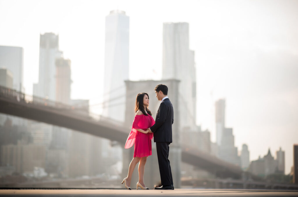 A couple stands closely facing each other on a rooftop with a soft-focus cityscape in the background, the woman in a vibrant pink dress and the man in a classic suit.