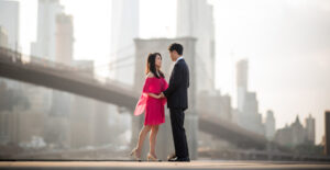 A couple stands closely facing each other on a rooftop with a soft-focus cityscape in the background, the woman in a vibrant pink dress and the man in a classic suit.