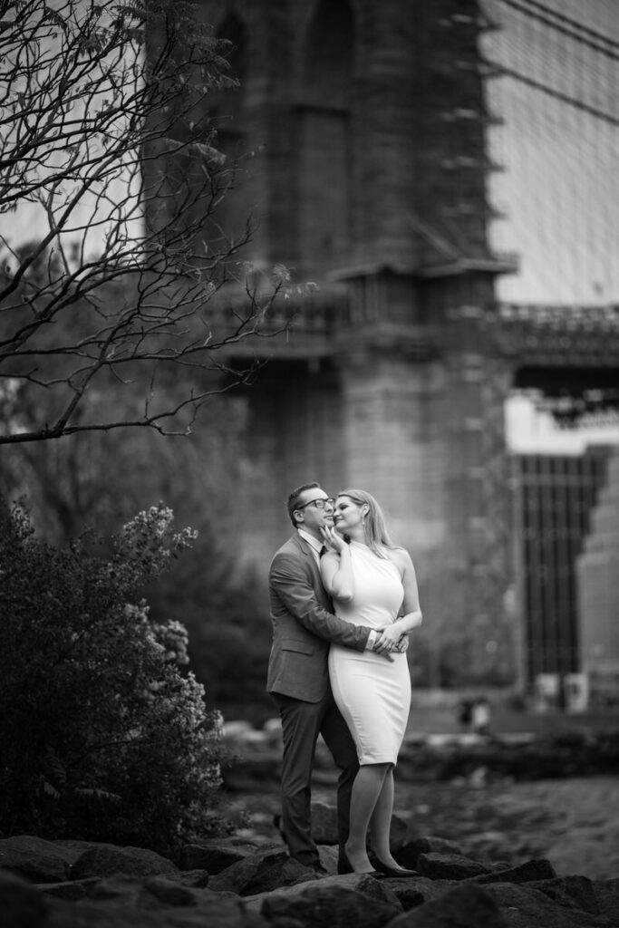 "A couple in a tender pose by a riverbank with the Brooklyn Bridge in the background, captured in black and white, reflecting a moment of affection