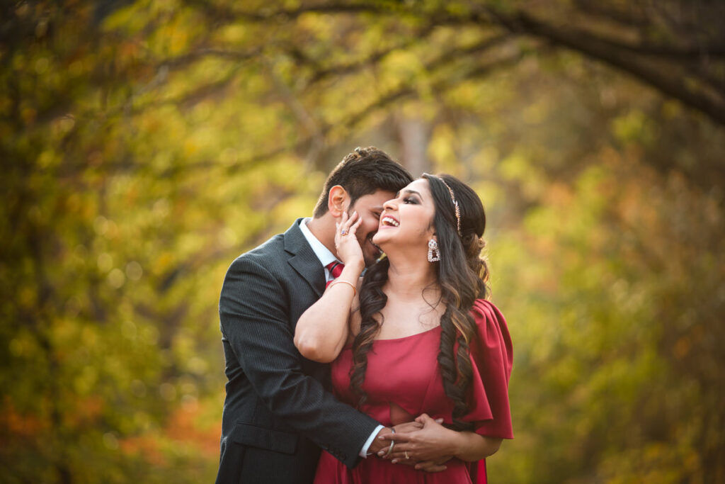 A man whispers into the ear of a woman who is laughing heartily, both dressed in formal attire amidst a lush autumnal setting, capturing a moment of joy and intimacy.