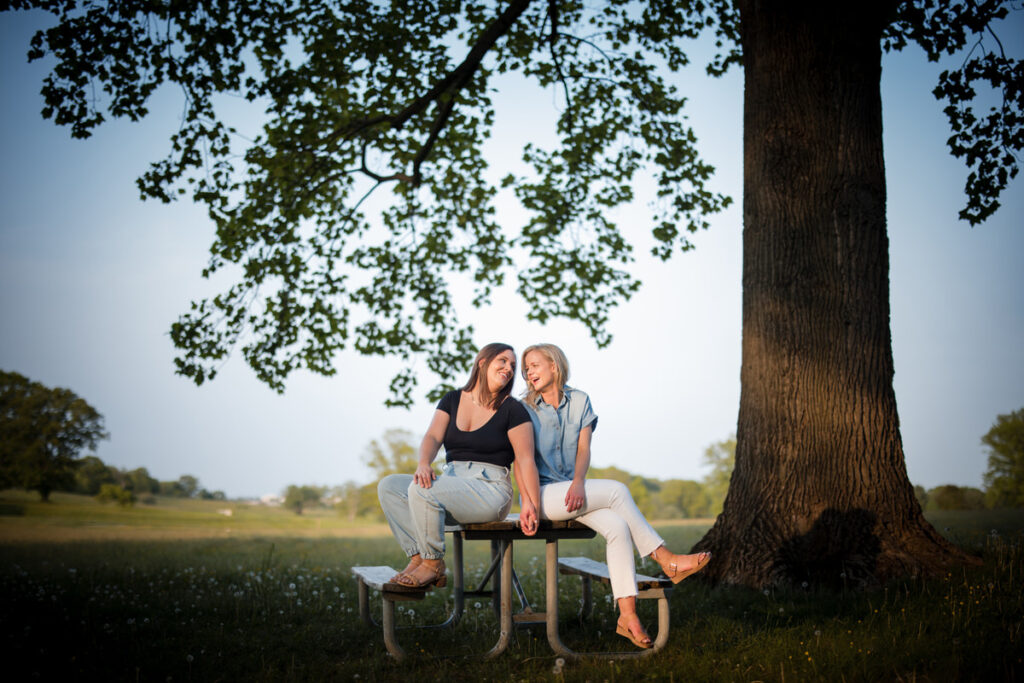 A couple sitting on a picnic bench under a large tree holding hands.