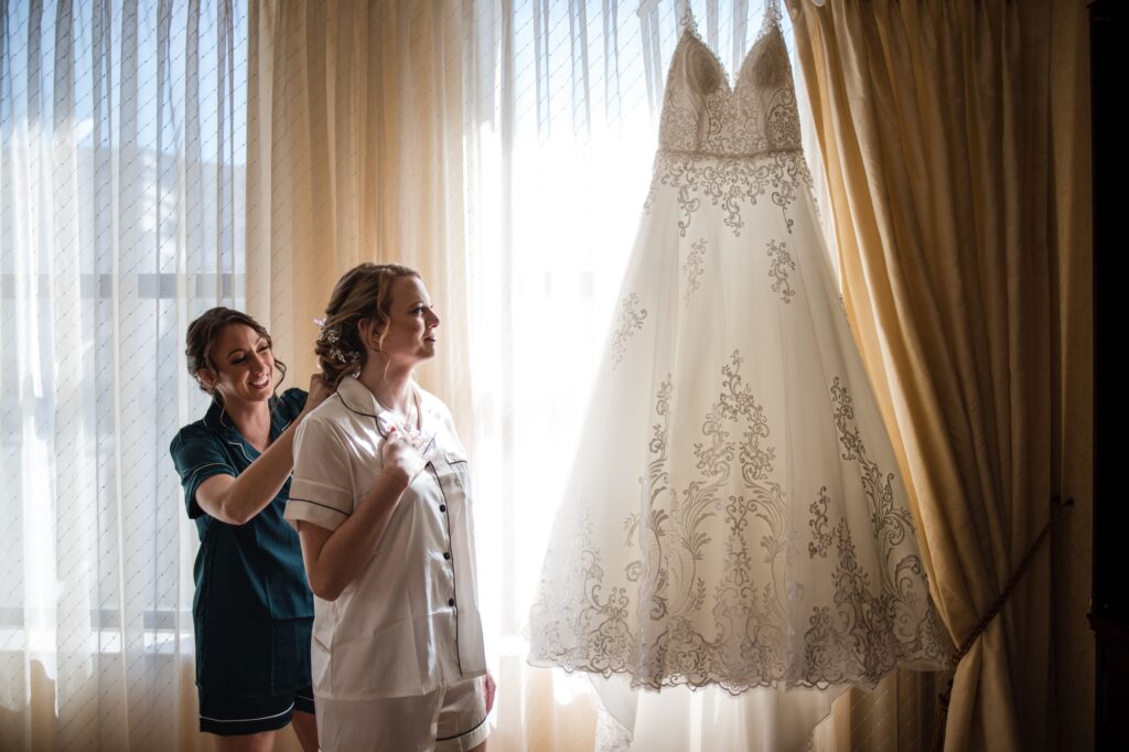 A bride is putting on her wedding dress in front of a window at the Hotel duPont wedding.
