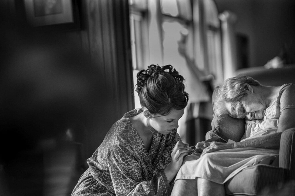 A black and white image capturing an intimate moment where a young woman in a patterned dress comforts an older woman dozing in a chair, highlighting a tender and caring interaction.