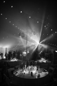 A monochrome image capturing the essence of a wedding celebration under a tent adorned with string lights, focusing on the guests and the ambiance of the sparkling venue.