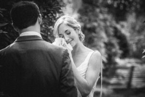 An emotional black and white photograph of a bride, gently wiping away tears with a tissue, standing opposite a man in a suit, suggesting a poignant moment during a wedding ceremony.