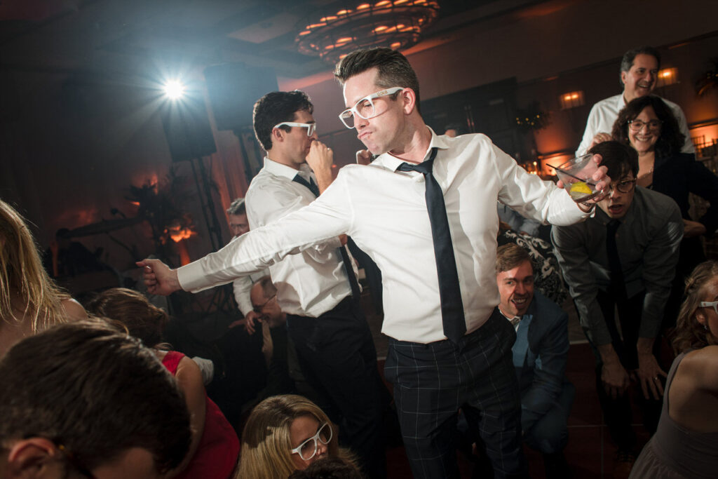 A lively black and white dance floor scene where a man in a white shirt and tie leads a dance, surrounded by guests in festive spirits at a wedding reception.