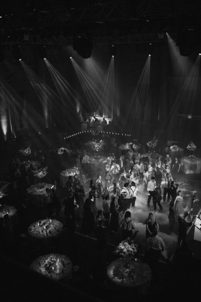 An atmospheric black and white image of a wedding reception held in a grand hall, with guests dancing under beams of light that create a dynamic and celebratory mood.