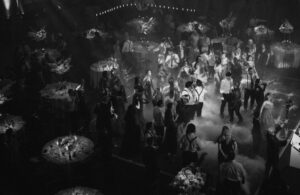 An atmospheric black and white image of a wedding reception held in a grand hall, with guests dancing under beams of light that create a dynamic and celebratory mood.