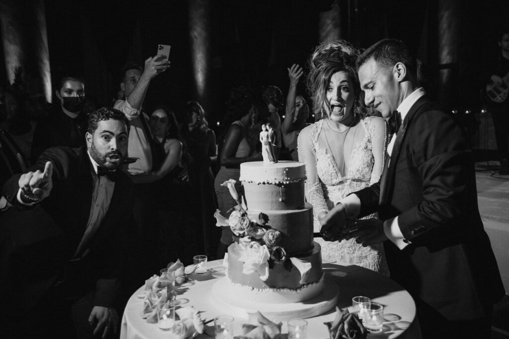 A couple cutting their wedding cake, the bride laughing heartily while the groom gestures playfully, with guests gathered around them in a dimly lit, candid celebration scene.
