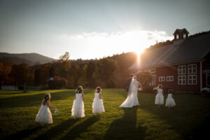 A serene outdoor scene of a bride and groom followed by four young flower girls in white dresses, walking across a sunlit grassy field towards a rustic red barn.