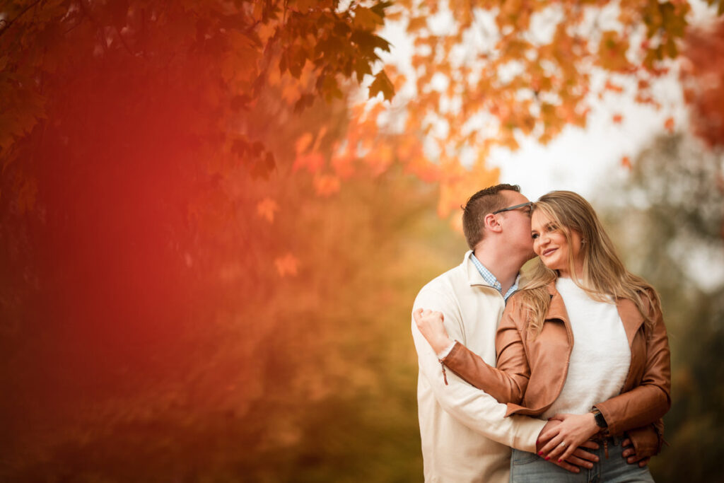 A couple in a warm embrace, surrounded by the brilliant reds and oranges of autumn leaves, with the man kissing the woman's forehead.