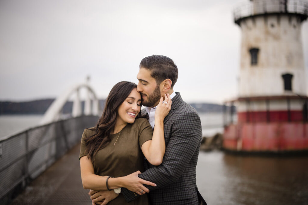A couple embracing on a walkway by the water, with the man kissing the woman's forehead as she smiles contently, and a lighthouse in the soft-focused background.