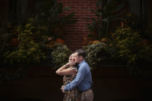 A man playfully covers a woman's eyes with his hand from behind, both smiling, in front of a brick wall adorned with lush greenery and autumn decorations.