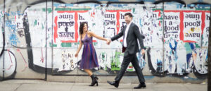 A couple walking hand in hand in front of a colorful urban graffiti wall, with the woman looking back and smiling at the camera.