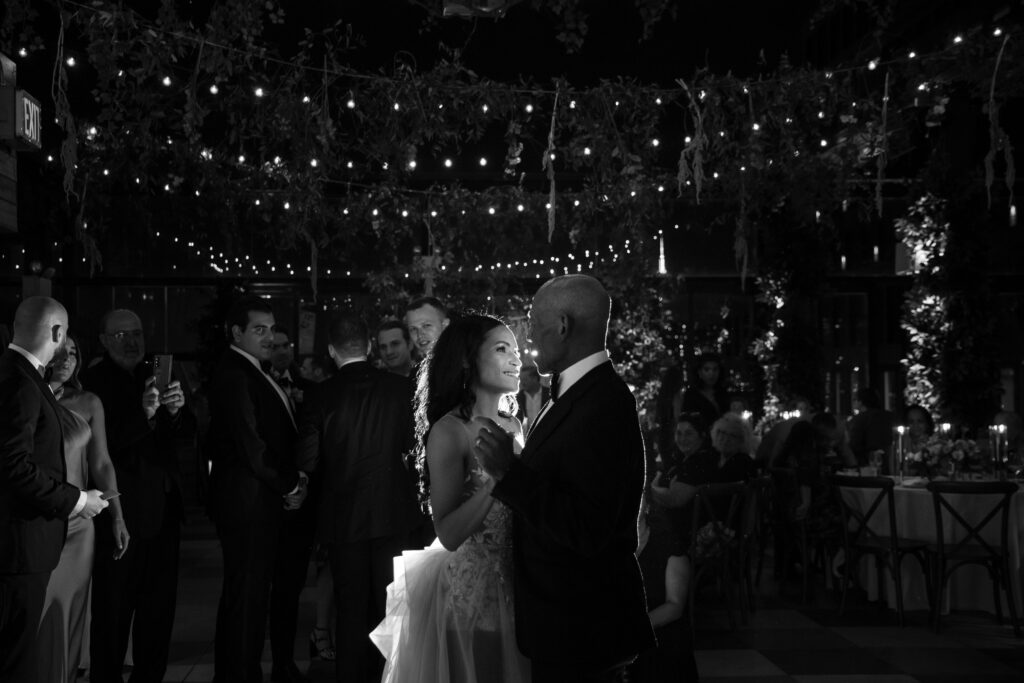 A couple shares a parent dance under string lights at an evening wedding on the 74 Wythe rooftop while surrounded by guests.