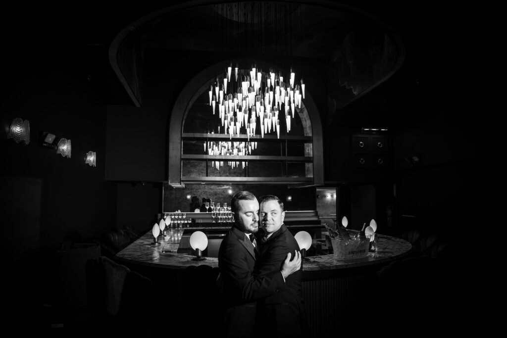Two grooms embracing at a dimly-lit bar with a decorative chandelier overhead, celebrating their 74 Wythe rooftop wedding.