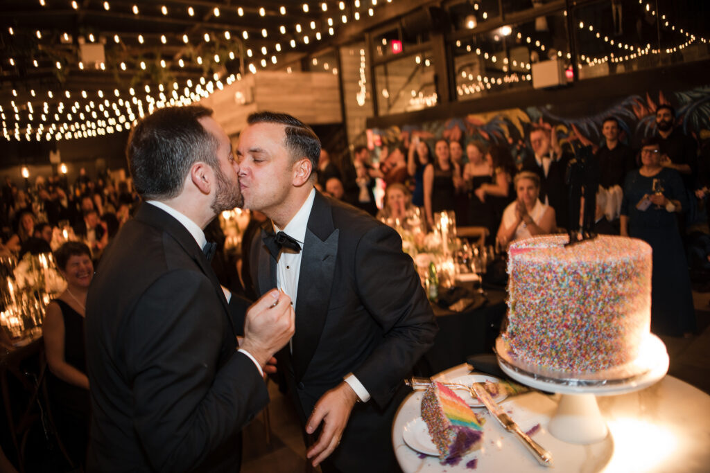 Two grooms in tuxedos kissing at their rooftop wedding at 74 Wythe, with a cake in the foreground and onlookers in the background.