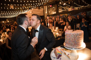 Two grooms in tuxedos kissing at their rooftop wedding at 74 Wythe, with a cake in the foreground and onlookers in the background.