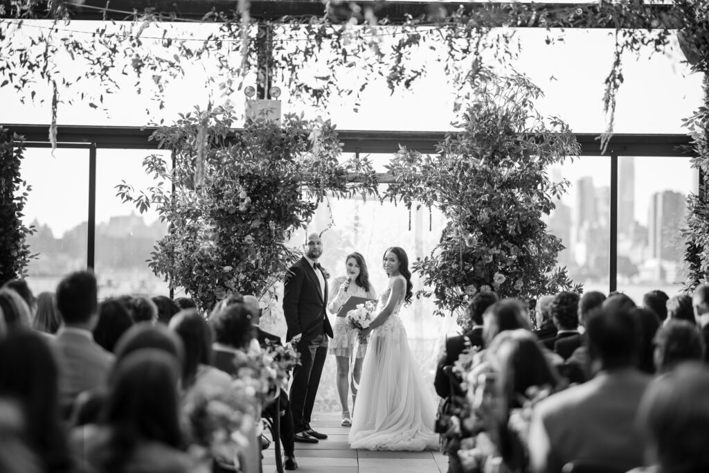 A black and white photograph of a bride and groom standing under a floral archway with guests seated on either side during a 74 Wythe rooftop wedding ceremony.