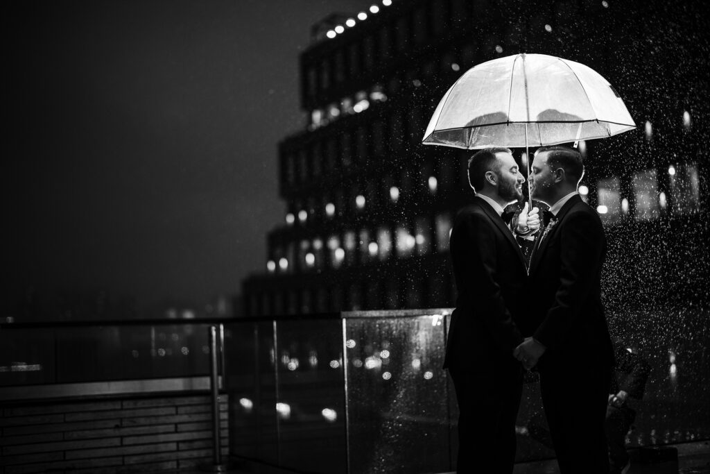 Two grooms sharing an intimate moment under an umbrella on a rainy evening at their 74 Wythe rooftop wedding.
