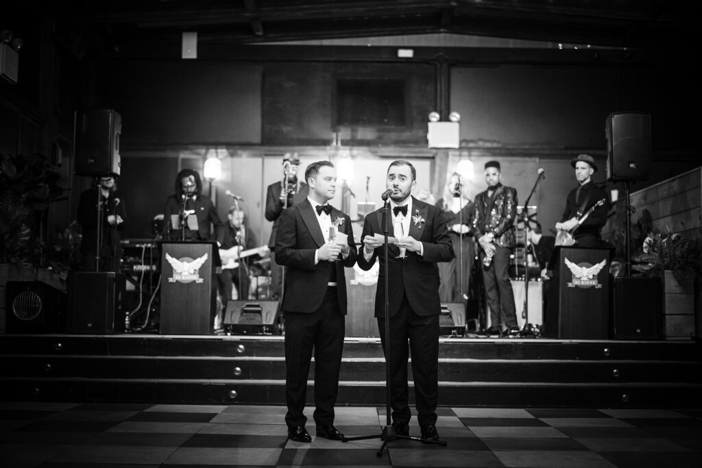 Two grooms in tuxedos holding microphones give a toast at a 74 Wythe rooftop wedding with a live band in the background.