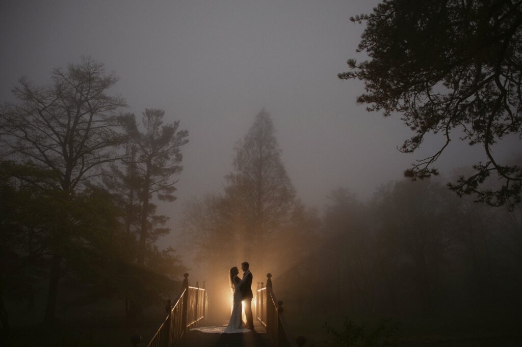 A couple stands on a bridge enveloped in fog with a gentle backlight at their Pleasantdale Chateau wedding.