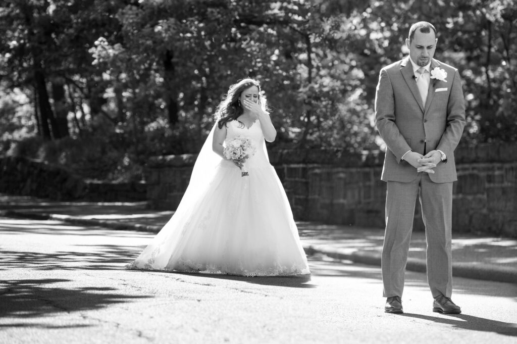 A bride and groom stand apart on a sunlit path at the Bronx Zoo Stone Mill wedding first look, both appearing emotional with the bride wiping a tear from her eye.