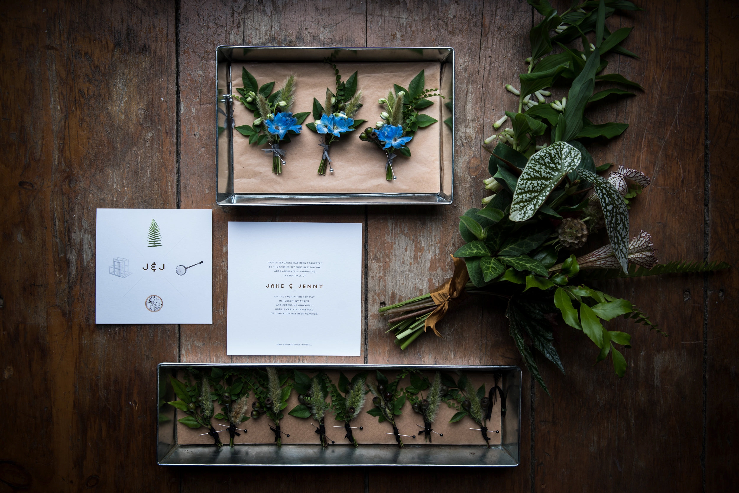 A Hudson Valley wedding invitation card alongside a boutonniere, a bouquet, and greenery on a wooden surface.