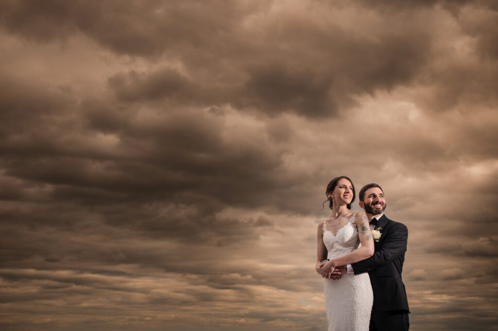 A bride and groom pose together under a dramatic cloudy sky at their Bourne Mansion wedding.
