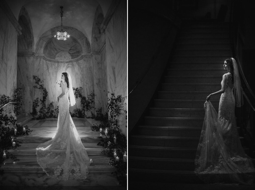 A bride in a lace gown stands elegantly at the bottom of a grand staircase at the Ritz Carlton Philadelphia wedding, surrounded by floral decorations in a black and white photograph.