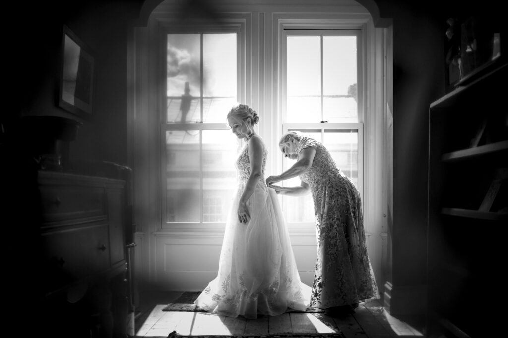 A bride in a white gown stands by a window while another individual helps adjust her dress in a room filled with natural light, at the Meadow Ridge on the Hudson Wedding venue.