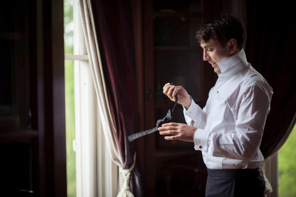 A groom in a white shirt and bow tie preparing for Hudson Valley Weddings at The Hill by a window.